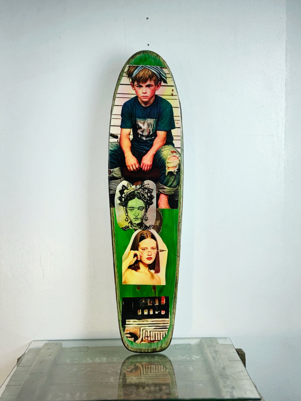 Skateboard 2 by David Bromley — portrait and seated-boy imagery on a green-toned skateboard deck, painted deck displayed above a glass-topped wooden crate.