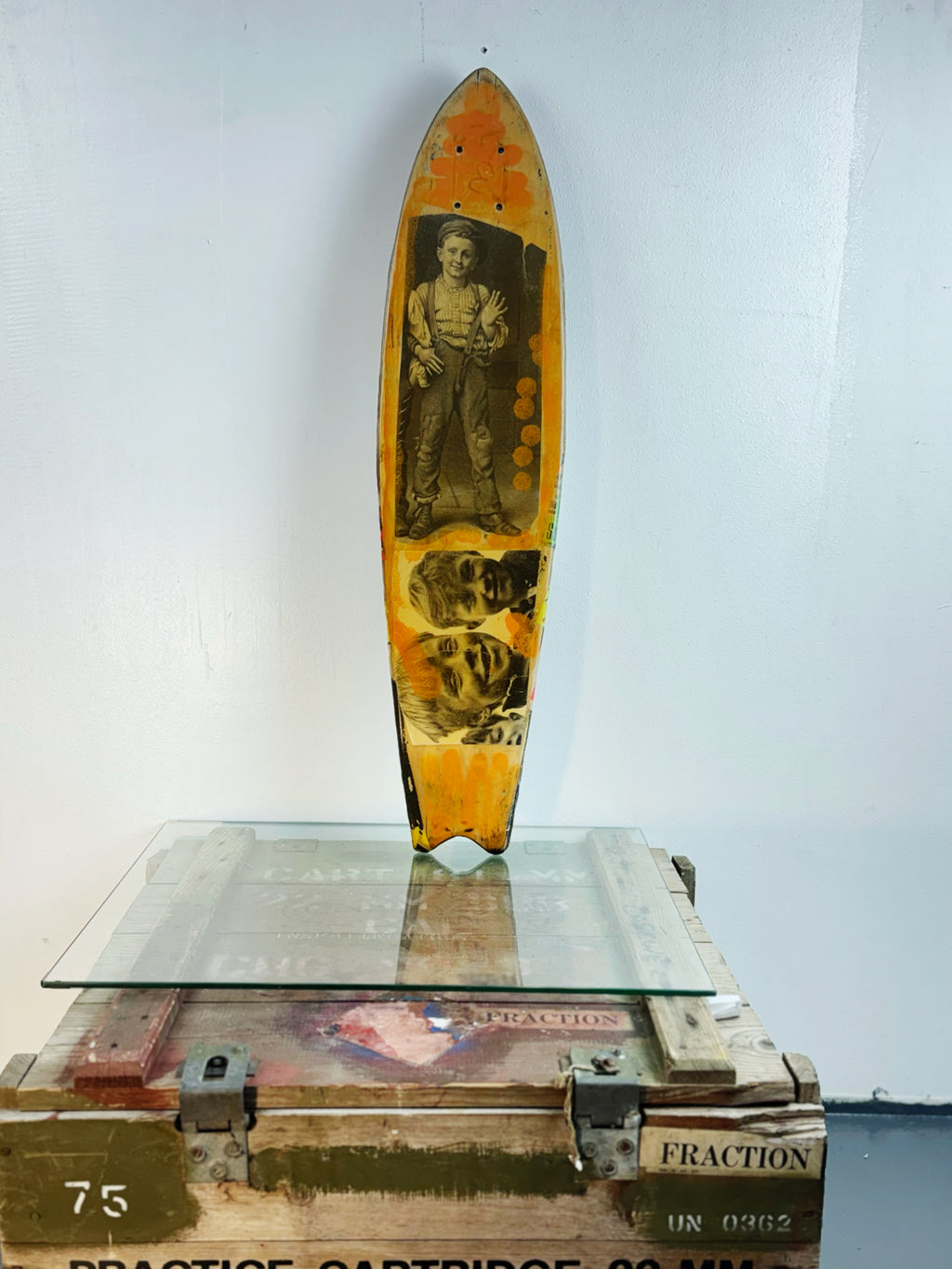 Skateboard 8 by David Bromley — bold poster lettering and yellow figurative imagery on a shaped skateboard deck, painted deck displayed above a glass-topped wooden crate.