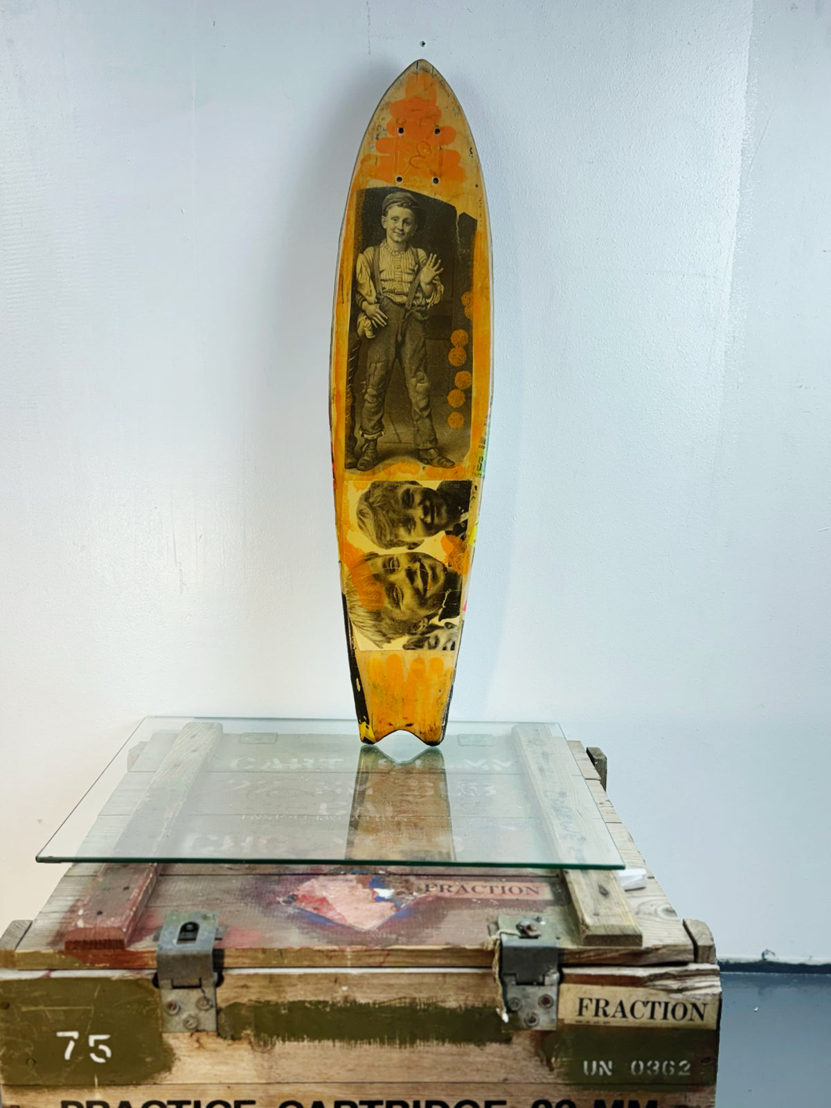 Skateboard 8 by David Bromley — bold poster lettering and yellow figurative imagery on a shaped skateboard deck, painted deck displayed above a glass-topped wooden crate.