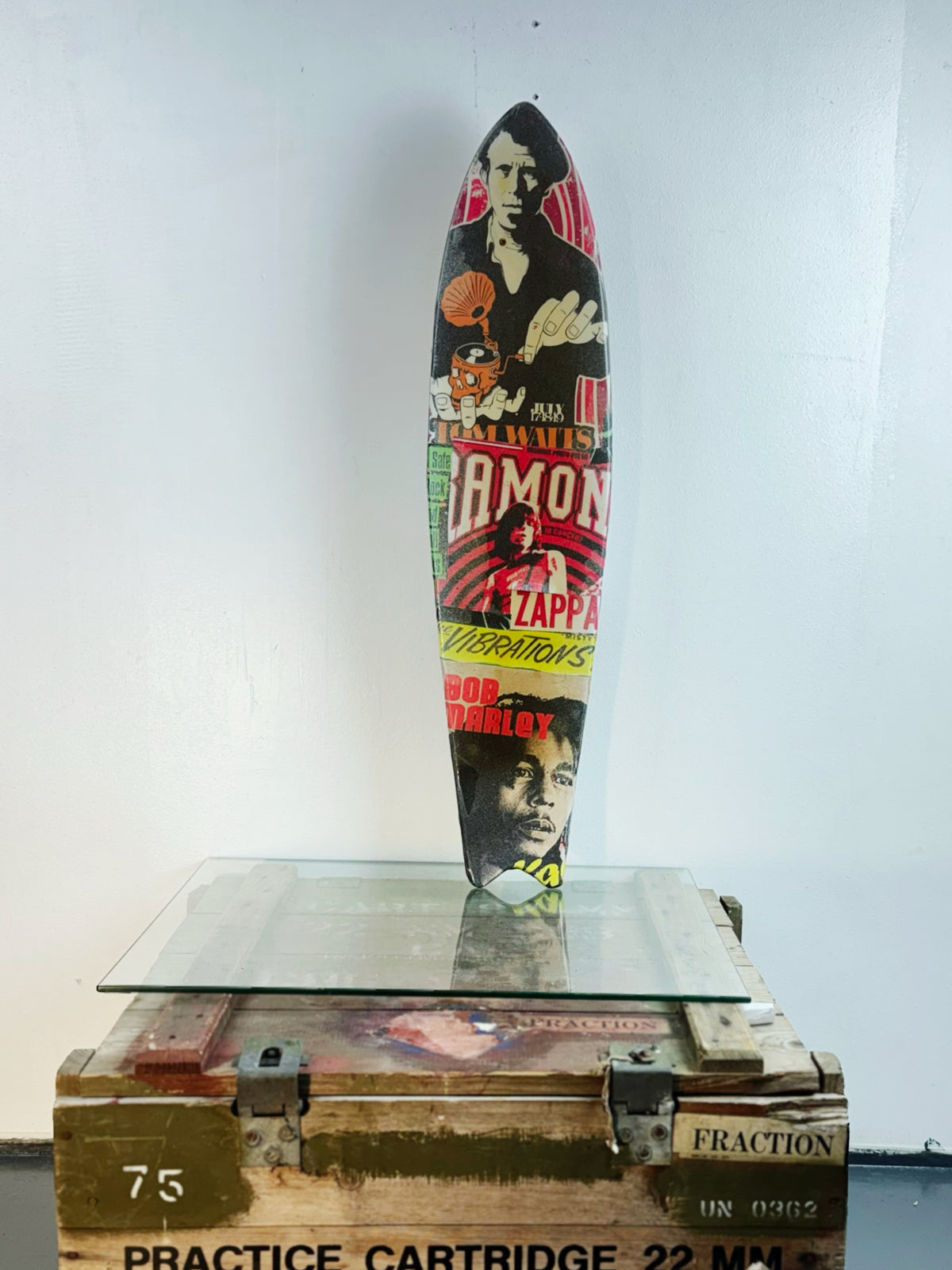 Skateboard 8 by David Bromley — bold poster lettering and yellow figurative imagery on a shaped skateboard deck, painted deck displayed above a glass-topped wooden crate.