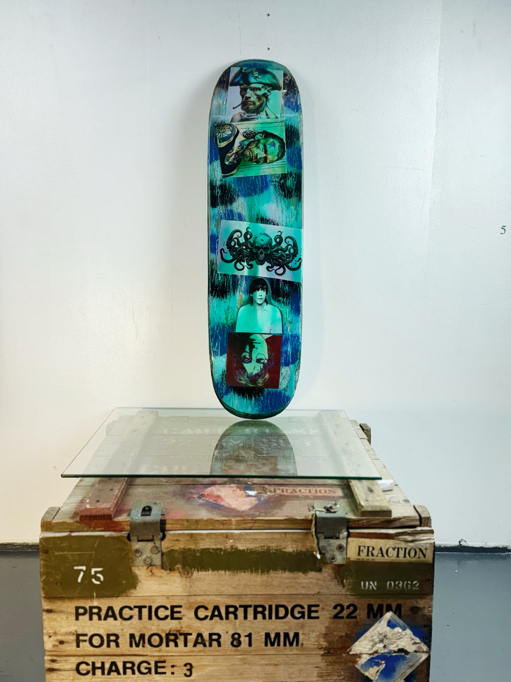 Skateboard 3 by David Bromley — blue-green collage and colourful portraits on a skateboard deck, painted deck displayed above a glass-topped wooden crate.