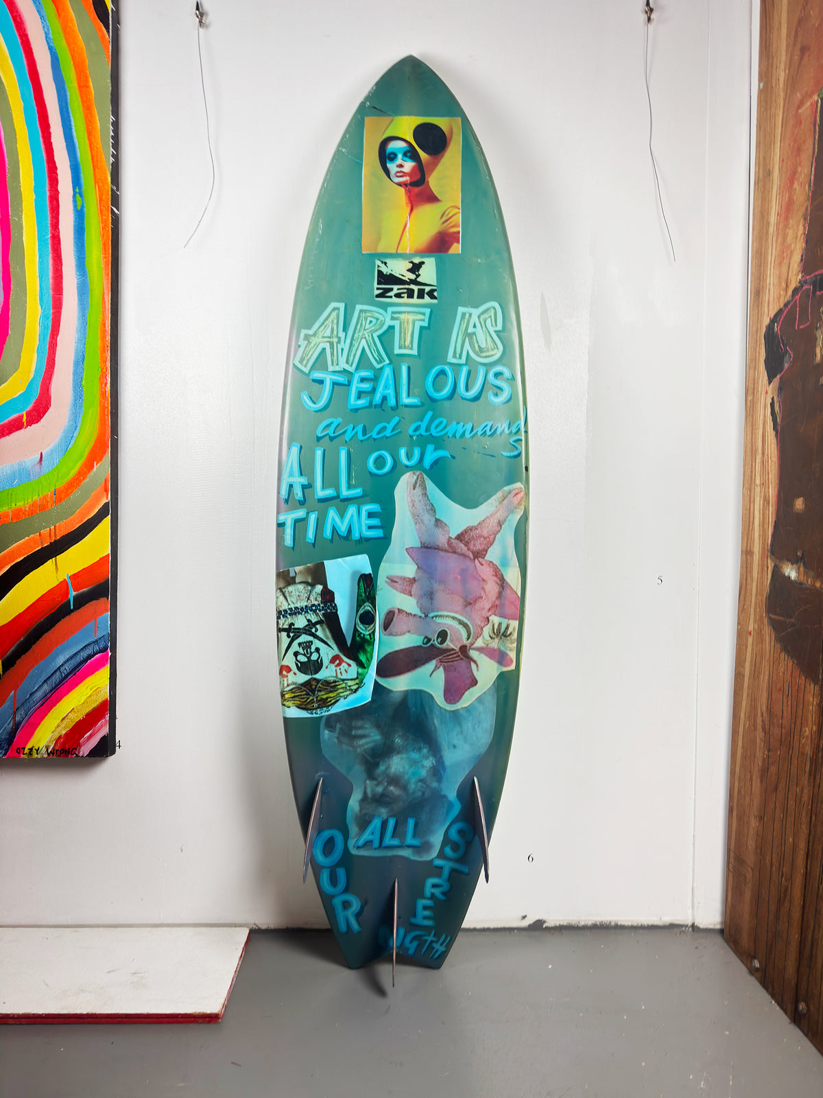 Blue Skulls Surfboard by David Bromley — turquoise lettered side standing in the gallery.