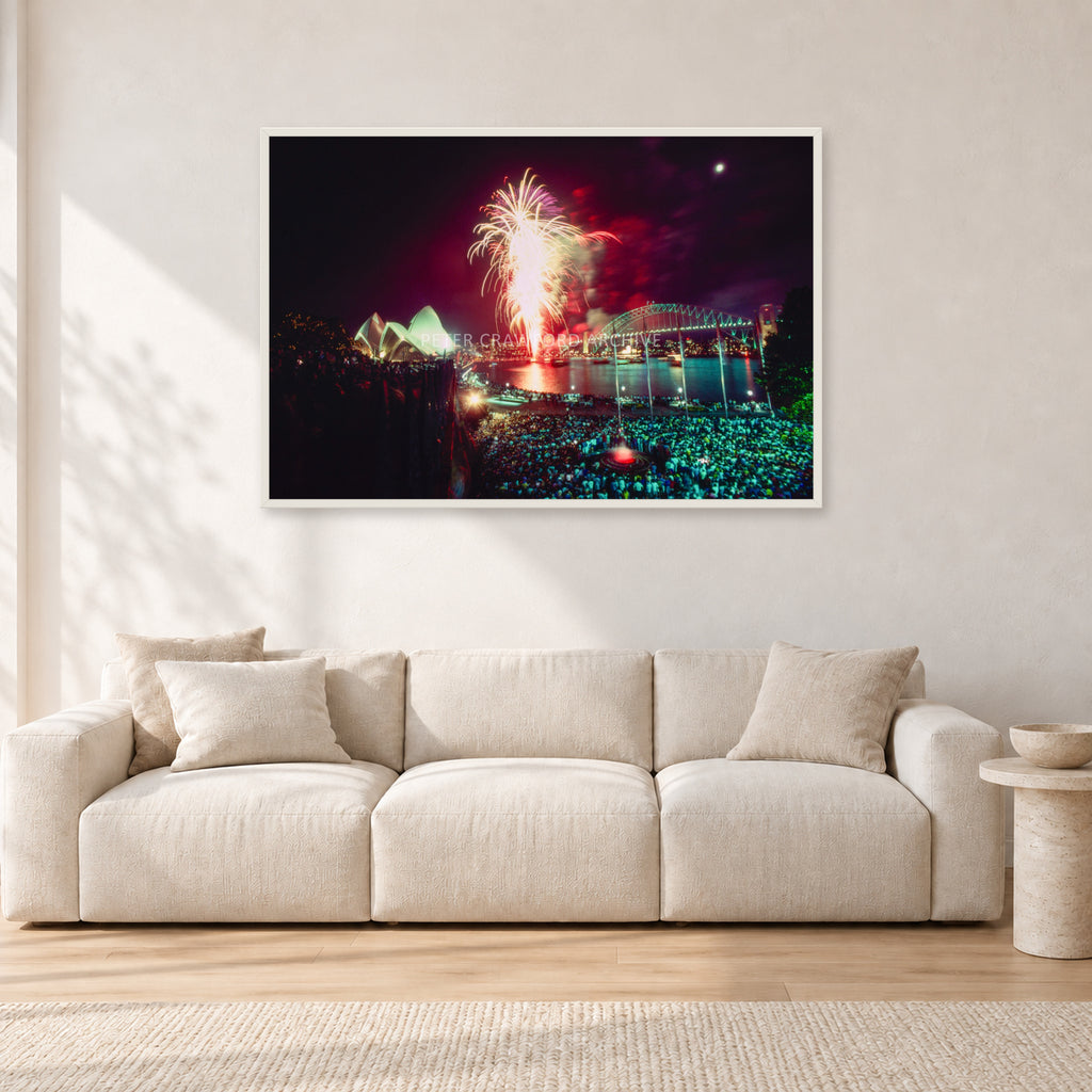 Tom Carroll and Aaron Chang, Waimea 1985 by Peter Crawford — 120 × 80 cm, white frame, shown in a light minimalist living room.